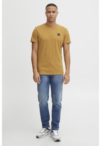 T-shirt kratkih rukava boje senfa s crnim logom, u kombinaciji s svijetloplavim trapericama i sivim tenisicama s mornarsko plavim detaljima.