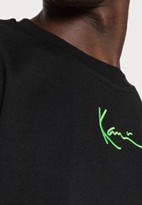 Černé bavlněné tričko s klasickým výstřihem, na levé straně je výrazný vyšívaný podpis v neonově zelené barvě. Hladká textura, volný střih.