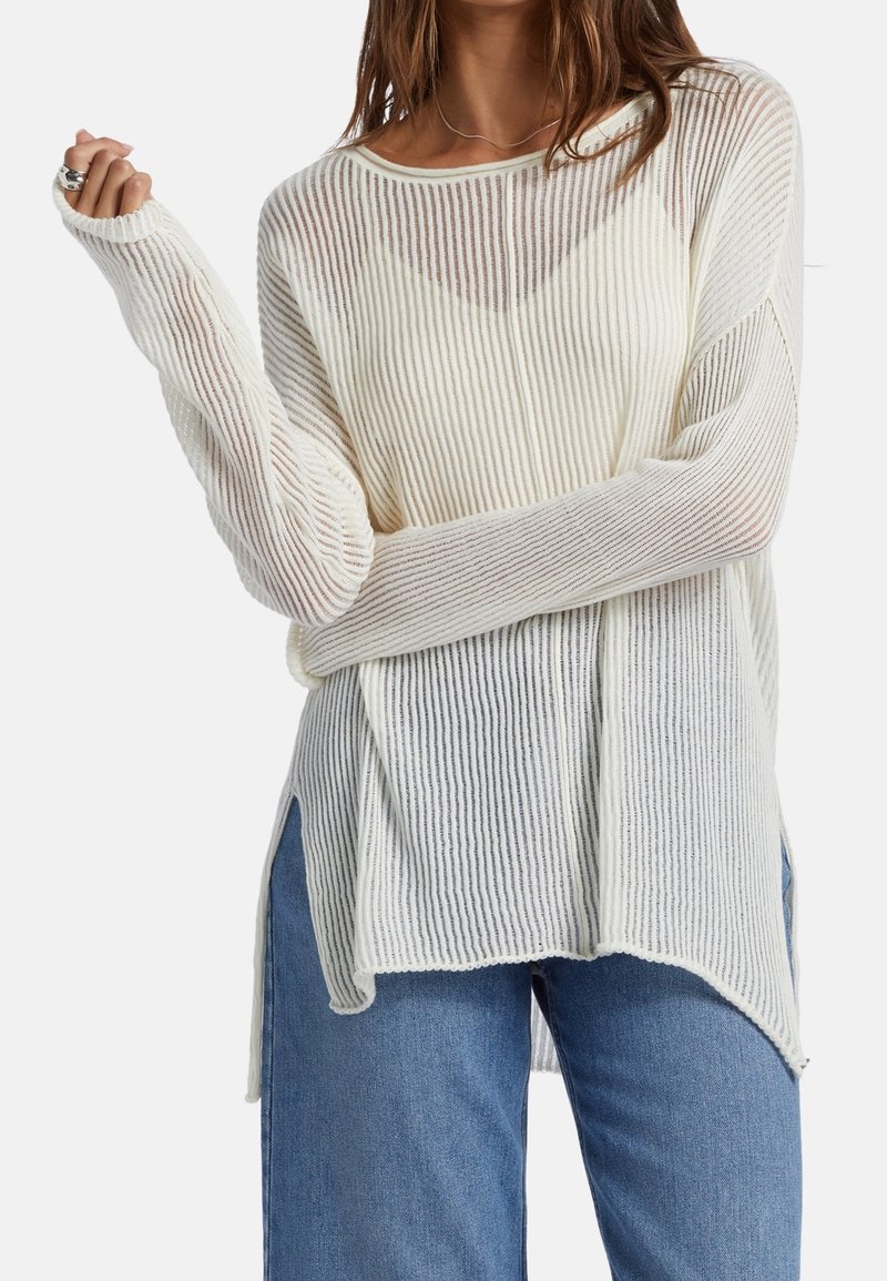 Rippstrickpullover in Off-White mit langen Ärmeln, weitem Ausschnitt, seitlichen Schlitzen und strukturiertem Muster, kombiniert mit blauen Jeans.