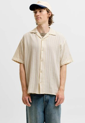 Ung mand iført en beige kortærmet skjorte med knapper, blå jeans og en hvid og marineblå baseballkasket, stående mod en ensfarvet baggrund.