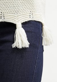 Haut tricoté couleur crème avec un motif texturé et des franges à la base, associé à un denim bleu foncé.