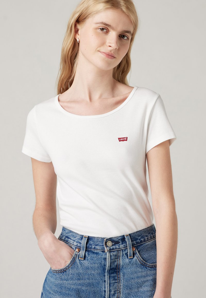 Camiseta blanca de cuello redondo en algodón suave con mangas cortas. Presenta un pequeño logo rojo de Levi's en el pecho. Combinada con unos vaqueros azules de cintura alta.