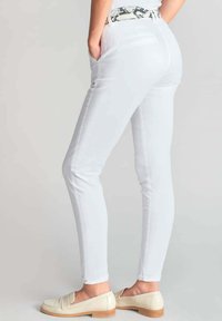 Weiße, schmale Fit-Hosen mit Fronttaschen, versehen mit einem Schlangenprint-Gürtel. Der Stoff wirkt glatt und dehnbar, mit einer sauberen, taillierten Silhouette.