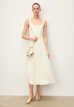 Femme en robe midi blanche sans manches tenant une petite pochette métallique dorée avec une chaîne, portant des sandales dorées à talons blocs.