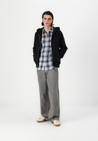 Schwarze Zip-Up-Hoodie über einem blau-weißen Karoshemd, kombiniert mit grauen gestreiften Weitbeinen und weißen Sneakers mit braunen Sohlen.