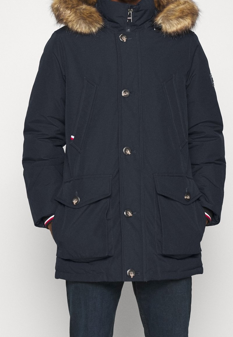 Tmavě modrý parka kabát s kapucí lemovanou umělou kožešinou, velkými předními kapsami a zapínáním na patentní knoflíky. Má prošívanou texturu a kontrastní červené a bílé detaily.