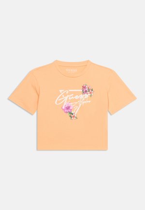 Kuttet T-skjorte i ferskenfarge med hvit "Guess Los Angeles"-logo inne i en trekant, dekorert med rosa og oransje tropiske blomster.