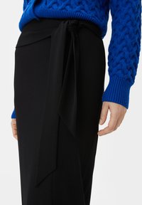 Zwarte high-waisted rok met een strikdetail aan de zijkant, gecombineerd met een gestructureerde blauwe gebreide trui. De rok heeft een gladde stofafwerking.