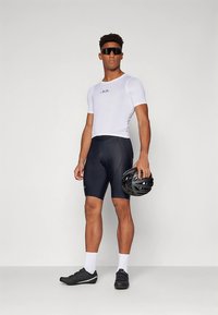 Fehér rövid ujjú kerékpáros mez, fekete párnázott rövidnadrág, fekete kerékpáros cipő, fekete sisakot tartva, világos háttér előtt áll.