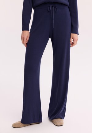 Granatowe spodnie o szerokich nogawkach z sznurkiem w pasie, noszone z dopasowanym granatowym swetrem i beżowymi teksturowanymi butami wsuwanymi.