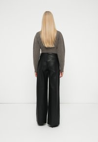 Vrouw met lang blond haar, gekleed in een bruine trui en hoge wijde zwarte leren broek, die met haar rug naar de camera staat tegen een effen achtergrond.