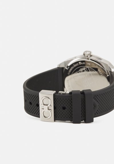 Mustast kummist käekellarihm perforatsiooniga tekstuuriga, hõbedane metallist pandla logoga ja roostevabast terasest korpus nähtav küljelt.