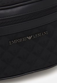 Černá taška s texturou a hladkým koženým povrchem, výrazným zipem a vyrytým kovovým logem "EMPORIO ARMANI".