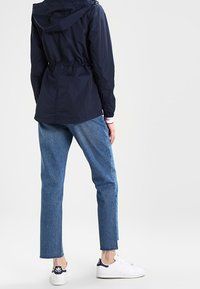 Giacca leggera blu navy con vita elasticizzata e cordoncino, maniche lunghe e cappuccio, abbinata a jeans in denim blu tagliati e sneakers bianche.