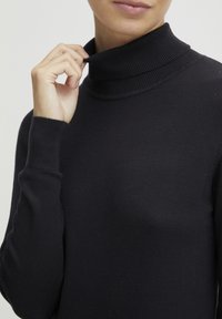 Eine Person, die den Kragen eines schwarzen gerippten Rollkragenspullis anpasst, zeigt dabei Details des Oberkörpers und der Hände.