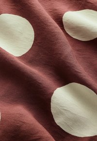 Texturovaná tkanina s velkými béžovými kruhy na rezavém pozadí, s viditelnou strukturou tkaní a měkkým, splývajícím vzhledem.