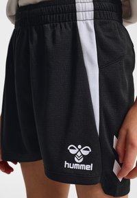 Schwarze Sportshorts mit weißen Seitenstreifen und Hummel-Logo am unteren Bein, getragen von einer Person, deren eine Hand in der Nähe des Saums sichtbar ist.