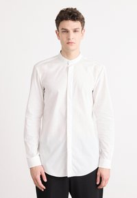 Camisa blanca de botonadura con un collar alzado; tejido de algodón suave; mangas largas; ligero brillo; dobladillo redondeado; diseño minimalista.