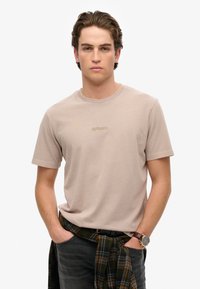 Beige t-shirt med korte ærmer og rund hals, med "Superdry" logo i lys broderi. Kombineret med mørke jeans og en ternet skjorte knyttet om taljen.