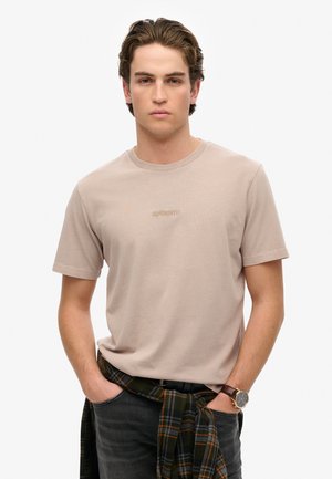 T-shirt beige à manches courtes avec un col rond, arborant le logo "Superdry" en broderie clair. Associé à un jean foncé et une chemise à carreaux nouée à la taille.