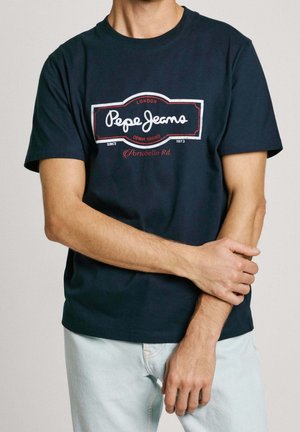 Uomo che indossa una t-shirt Pepe Jeans blu navy con logo bianco e rosso, che tiene il braccio sinistro con la mano destra, vestito con jeans chiari.