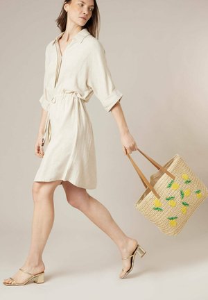 Vrouw in een beige overhemdjurk die loopt, met een strooien draagtas met gele bloemmotieven en gouden sandalen met blokhak.