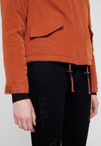 Veste orange rouille avec grande poche avant et poignets à boutons-pression portée avec jean noir déchiré, montrant la taille et la main gauche.