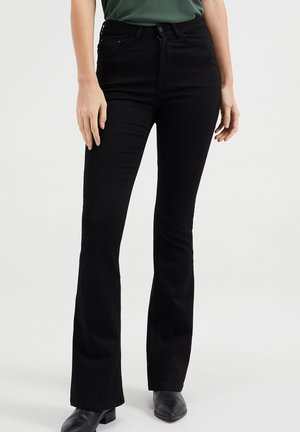 Schwarze, ausgestellt geschnittene Jeans mit glatter Textur, hoher Taille, Vordertaschen und Reißverschluss; kombiniert mit schwarzen Stiefeletten.