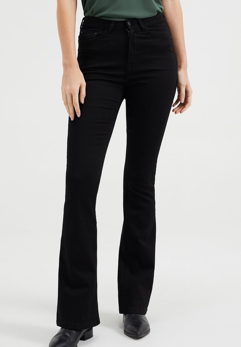 Black flared jeans with a smooth texture, high waist, front pockets, and a zipper fly; paired with black ankle boots.