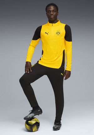 Sort og gul atletisk træningsdragt med lynlåskrave, der har tekstureret stof, Puma-logo og sorte fodboldstøvler på en sort og gul bold.