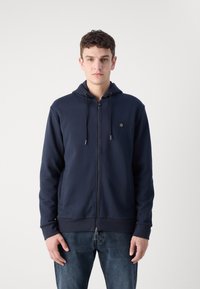 Navy zip-up hoodie van gladde stof met een voorkeurszak en een capuchon met koord. Heeft een klein logo op de borst. Er worden spijkerbroeken gedragen.