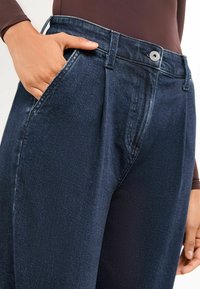 Temno modri denim hlače z visokim pasom in strukturiranim dizajnom, z širokimi nogami, sprednjimi gubami in stranskimi žepi. Vidna srebrna zgornja gumb.