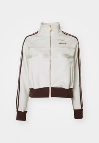 Vitt zip-up träningsjacka med bruna ribbade manschetter och nederkant, med glansiga randdetaljer och en broderad logotyp på bröstet.