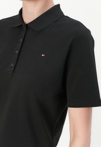 Černé polo tričko vyrobené z texturované látky s límečkem, tři knoflíkovou légu a malým vyšitým logem v červené, bílé a modré barvě.