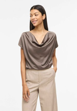 Kobieta o długich ciemnych włosach, ubrana w taupe satynową bluzkę z drapowaniem i beżowe spodnie z wysokim stanem, z rękami w kieszeniach, uśmiechająca się i patrząca w lewo.