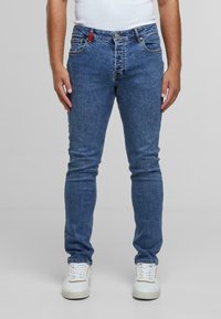 Kék farmernadrág slim fit szabással, öt zsebbel, gombos záródással és piros címkével. Fehér sportcipővel viselve. A textúra sima és egyenletes.
