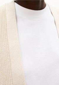 Beige pletený kardigan a bílý bavlněný tričko, které ukazuje směs různých textur s kulatým výstřihem a otevřeným předním designem.