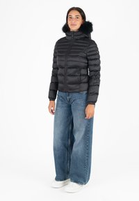 Giacca a vento nera con texture trapuntata e cappuccio foderato in pelo, abbinata a jeans in denim a gamba larga e sneakers bianche.