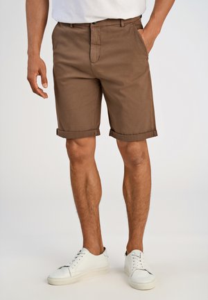 Shorts di cotone marroni con orli risvoltati, tasche frontali e zip. Abbinati a sneakers bianche. Sfondo neutro.