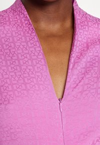 Primo piano di una persona di pelle scura che indossa una felpa viola con zip, decorata con un motivo a monogramma testurizzato e ripetuto.