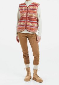 Kamizelka polarna w czerwone, pomarańczowe i niebieskie paski, zapinana na zamek; beżowa bluzka z długim rękawem; brązowe spodnie; beżowe botki na sznurowadła z teksturowaną podeszwą.