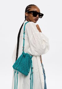 Borsa a secchiello all'uncinetto color teal con tracolla regolabile, caratterizzata da dettagli metallici e una finitura testurizzata. Indossata su una camicia bianca ricamata.
