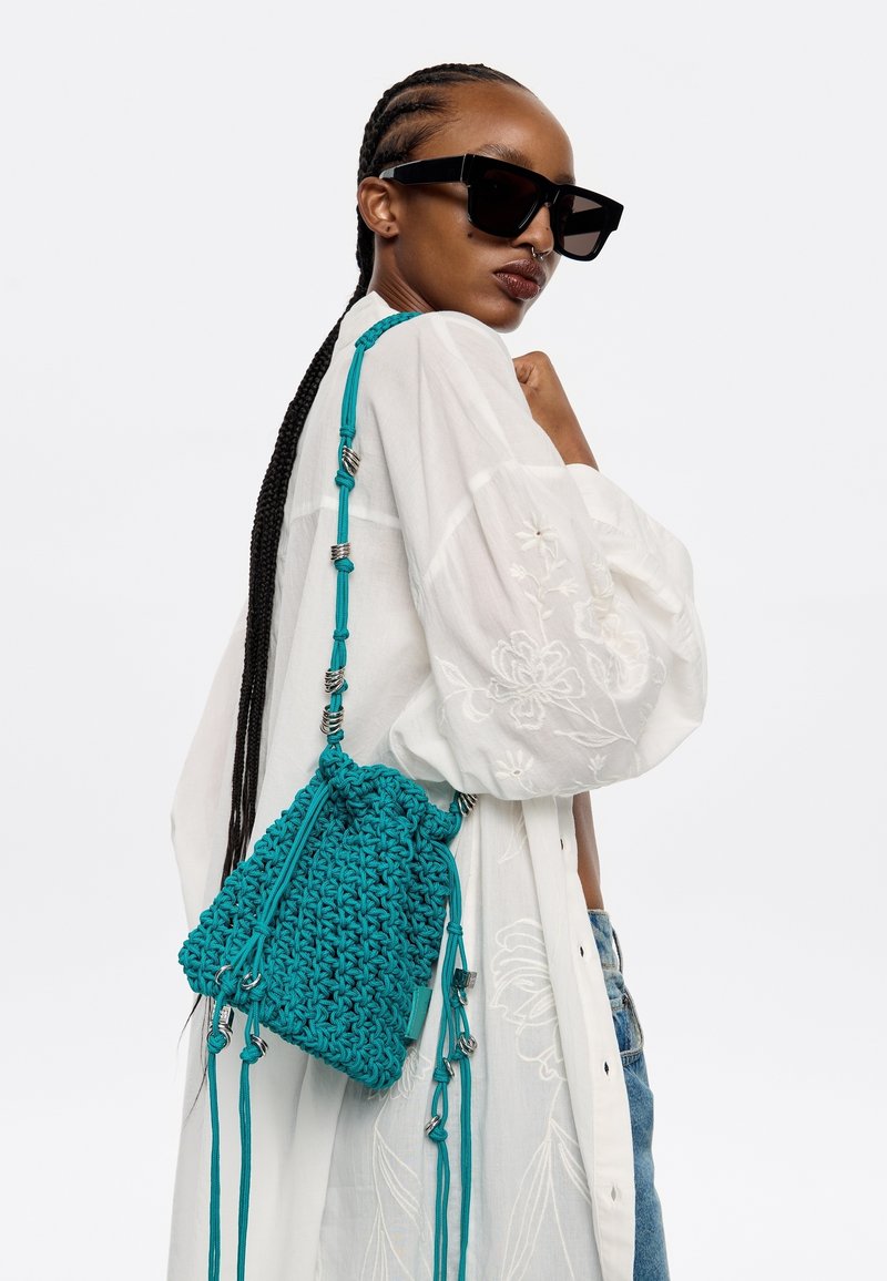 Borsa a secchiello all'uncinetto color teal con tracolla regolabile, caratterizzata da dettagli metallici e una finitura testurizzata. Indossata su una camicia bianca ricamata.