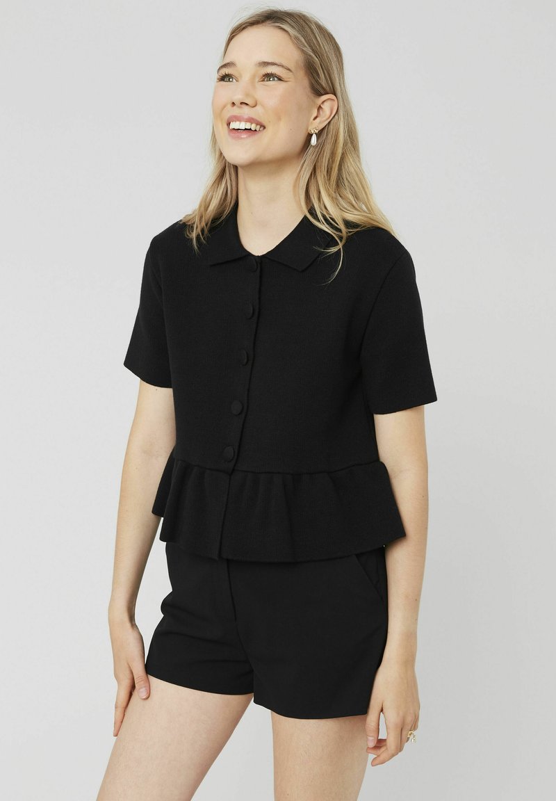 Haut noir à manches courtes avec un ourlet peplum, doté d'un col classique et de cinq boutons sur le devant. Texture lisse, porté avec un short assorti.