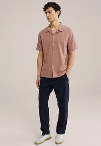 Camicia a maniche corte con colletto in rosa polveroso, realizzata in tessuto morbido, con design a bottone frontale. Abbinata a pantaloni blu navy e sneakers bianche.