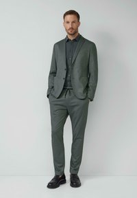 Mann steht mit den Händen in den Taschen, trägt einen grauen Anzug, ein schwarzes Hemd und schwarze Dressschuhe vor einem schlichten hellen Hintergrund.