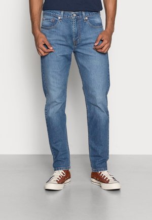 Mann trägt blaue Slim-Fit-Jeans und braune Low-Top-Sneaker mit weißen Schnürsenkeln, steht vor einem einfarbigen hellen Hintergrund.