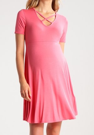 Donna che indossa un vestito premaman rosa al ginocchio con maniche corte e spalline incrociate a V, in piedi davanti a uno sfondo neutro.