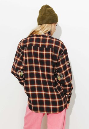 Chemise en flanelle noire à carreaux rouges et orange, ornée de motifs brodés de rayons de soleil sur les manches ; portée avec un pantalon en velours côtelé rose et un bonnet vert.