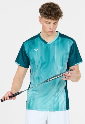 Ung mand iført en teal sportstrøje holder og inspicerer en sort badmintonketcher mod en ensfarvet baggrund.